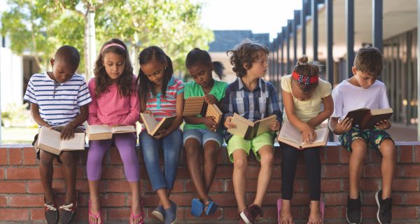 Front view of multi ethnic students reading book while sitting on brick wall at corridor in school