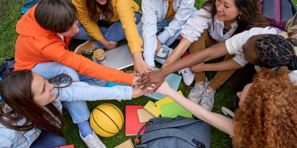 Top view portrait of multi-ethnic group young students joining hands in circle sitting on lawn of university campus. People gathered together enjoying break. Generation z and friendly relationships.
